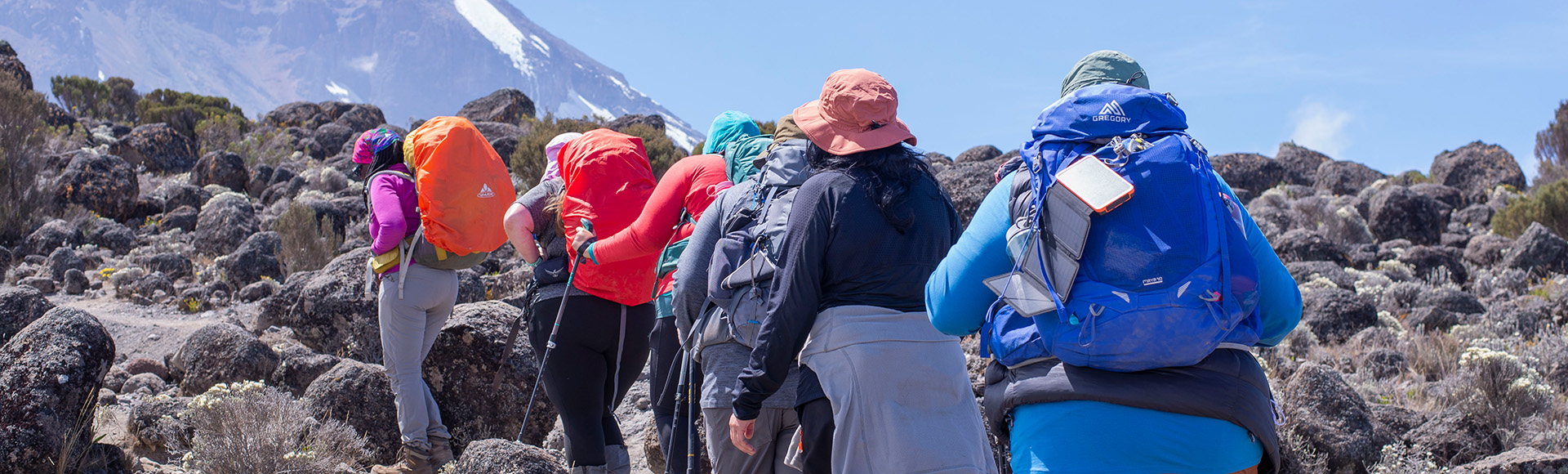 Trekkers with backpacks walking a rocky Kilimanjaro trail beneath the snow-capped summit
