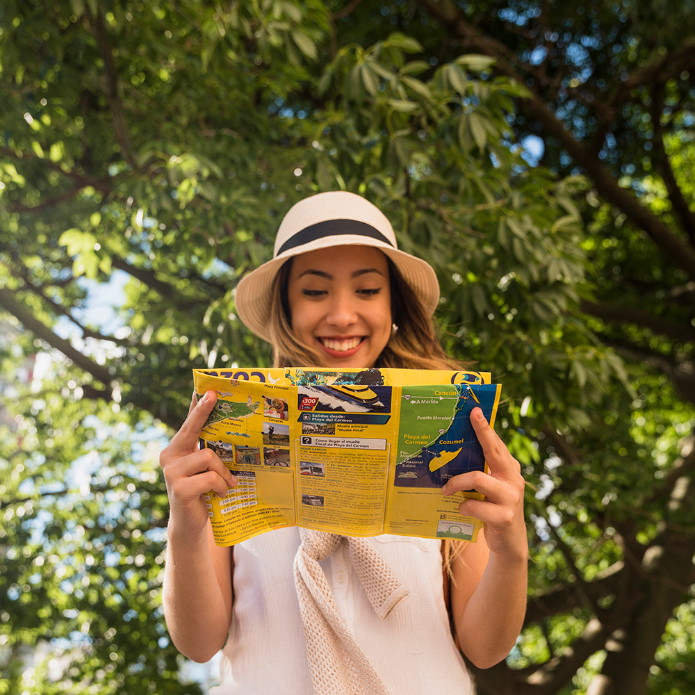 Smiling traveller in a sun hat reading a folded travel map under leafy trees